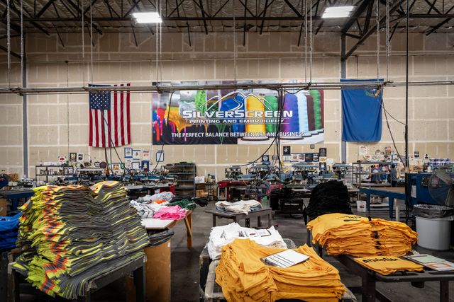 Screen printing shop floor with stacks of printed high-visibility and gold shirts in front of a Silverscreen Printing and Embroidery banner and an American flag